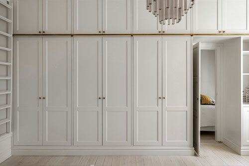 White built-in wardrobes with gold handles; one door is open, revealing a glimpse of a bedroom behind.