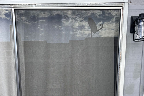 A window with a mesh screen reflects clouds and a satellite dish under a partly cloudy sky.
