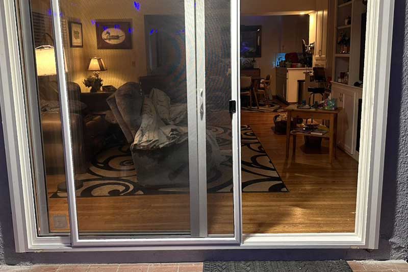 Glass sliding door reflects a living room with a couch, lamp, bookshelves, and a table with toys inside.